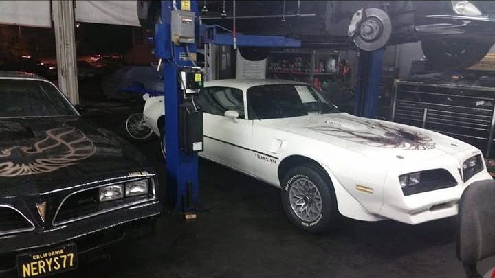 A black Pontiac Firebird Trans Am with the California plate NERYS77 beside a white Trans Am inside the workshop